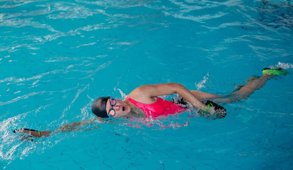 Female in pink swimwear swimming in blue water pool with hand paddle